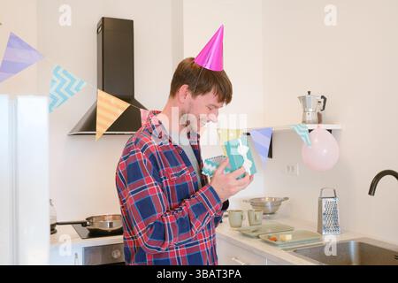 Young man receives gift at home, expressing surprise and happiness. Celebrating special occasion, he admires wrapped present in cozy, cheerful setting Stock Photo