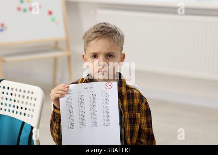 School grade. Sad boy holding answer sheet with letter C and minus symbol indoors Stock Photo