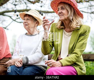 Two fashionable senior women relaxing on a bench during an outdoor gathering, drinking wine and sharing conversation. Elegant attire and natural smile Stock Photo