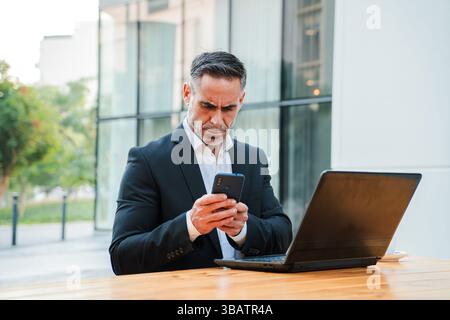 A serious businessman intently using his smartphone while simultaneously working on his laptop, showcasing a dedicated and professional approach to Stock Photo