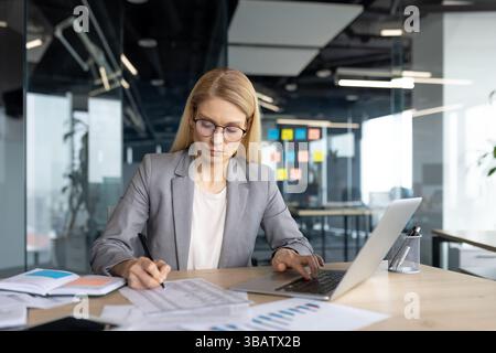 A businesswoman in a modern office setting is focused while working on a laptop and reviewing documents at her desk, analyzing data. Stock Photo