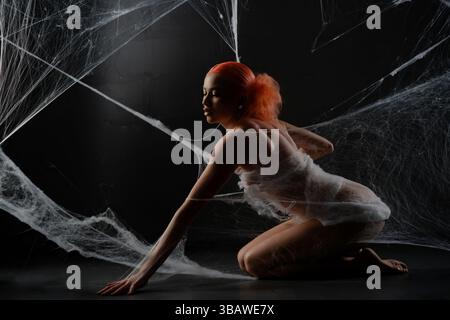 Red-haired woman trapped in a delicate web of white strands in dark setting Stock Photo