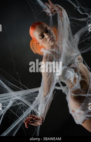 Red-haired woman entangled in an intricate net trap against a dark backdrop Stock Photo