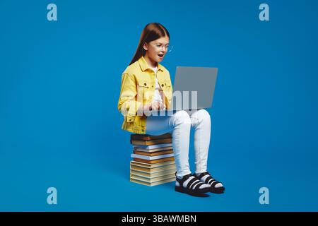 Surprised girl in yellow jacket using laptop while sitting on stack of books against blue background Stock Photo