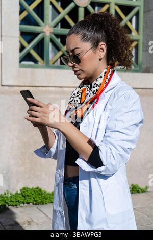 Young female doctor wearing white coat and sunglasses using mobile phone while standing outdoors Stock Photo