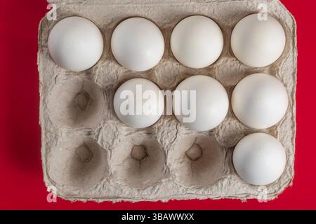 An open egg carton containing eight white eggs is placed on a vibrant red surface emphasizing color contrast and minimal arrangement. Stock Photo