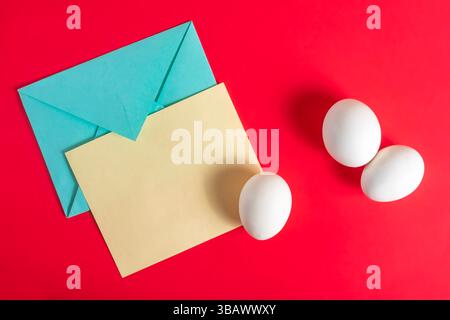 Top view, white eggs with blue envelope and yellow card on a red surface. Stock Photo