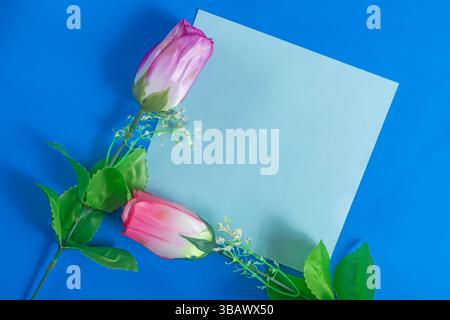 Pink artificial roses and a blank card on a vibrant blue background. Perfect for themes such as love, messages, invitations and celebrations. Stock Photo
