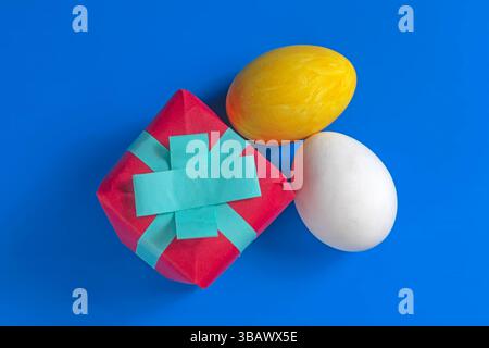 A red gift box with a blue ribbon and two colorful eggs, one yellow and one white, on a deep blue surface. Stock Photo
