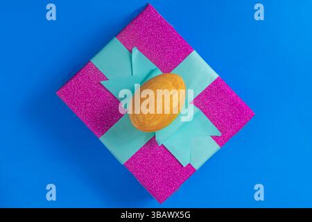 A shiny pink gift box with a turquoise ribbon and a golden egg on top, displayed against a blue background, symbolizes celebration and luxury. Stock Photo