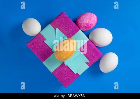 Vibrant Easter eggs decorated with a purple gift box decorated with blue ribbon, set against a bold blue background. Perfect for holiday, celebration Stock Photo