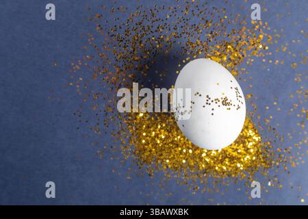 A white egg decorated with shimmering gold glitter placed on a vibrant blue background, symbolizing a happy Easter celebration. Perfect for spring and Stock Photo