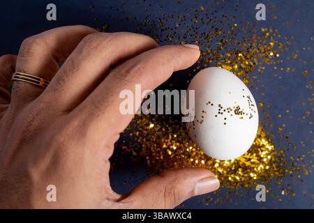 A close-up of a hand next to a white egg decorated with gold glitter, placed against a dark background decorated with shiny gold. Stock Photo