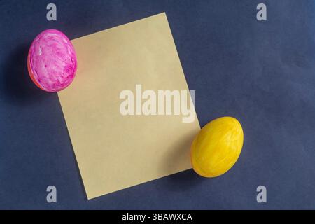 Brightly colored Easter eggs in pink and yellow next to plain yellow paper on a dark blue surface are spring celebrations. Stock Photo
