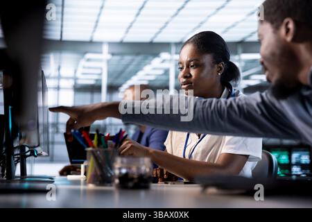 Data center engineer helping colleague, pointing finger to screen, using programming languages to construct algorithms and build applications. Server farm IT expert showing code to coworker on monitor Stock Photo