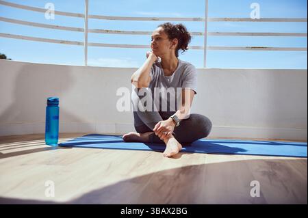Portrait of a woman seated on a yoga mat on a balcony. She's reflecting in a relaxed posture, epitomizing balance, tranquility, and mindfulness amidst Stock Photo