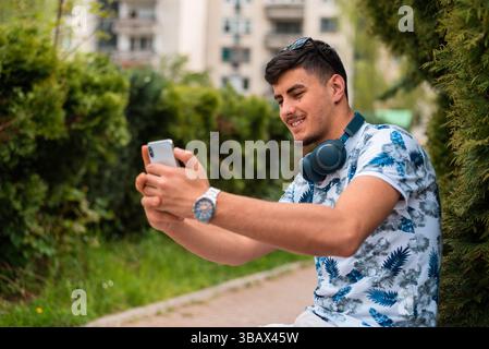 A young happy urban stylish man taking a selfie with a mobile phone in the city park. Stock Photo