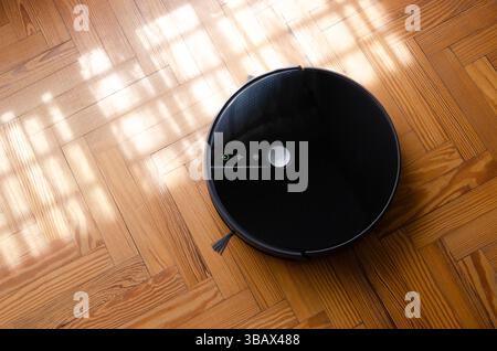 Black robot vacuum cleaner cleaning a wooden floor. Stock Photo