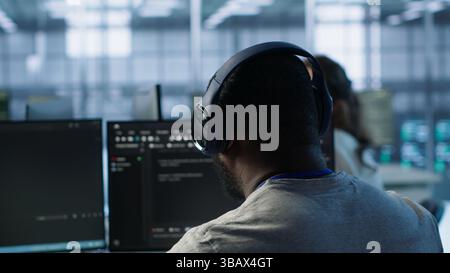 System administrator working in computer network security data center listening music. African american worker wearing headphones monitoring server racks energy consumption, camera A Stock Photo