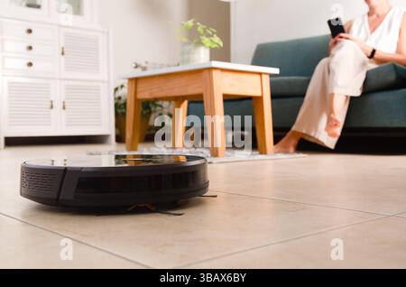 A robot vacuum cleaner cleaning the floor and a woman with mobile phone sitting on a sofa at home. Stock Photo