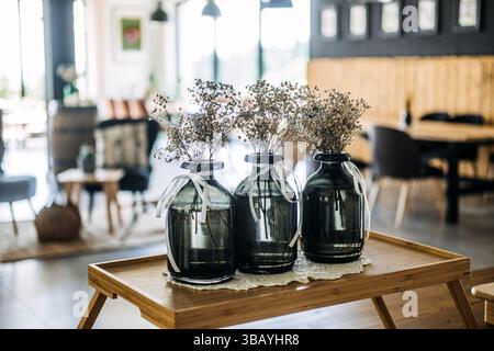 Three dark glass vases with dried flowers and white ribbons arranged on a wooden tray in a cozy cafe interior. Slow living, mindful design, interior Stock Photo