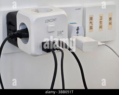 Multiple USB and plug adapters connected to a LENCT power hub mounted on a wall socket in a modern Chinese bathroom or living space. Stock Photo