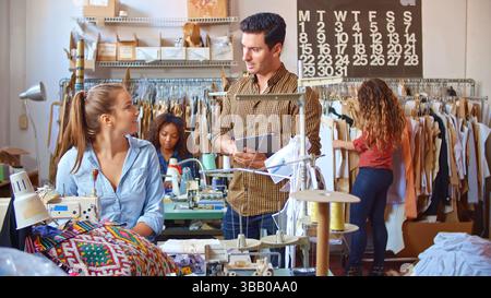 Male Supervisor With Digital Tablet Talking To Woman Using Sewing Machine In Clothing Factory Stock Photo