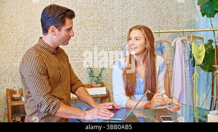 Male and Female Sales Assistants Using Laptop At Sales Desk Stock Photo