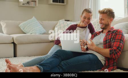 Young Couple Sitting On Floor In Lounge Using Laptop Together Stock Photo