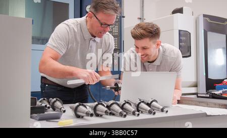 Male Engineer And Apprentice Measuring Components Whilst Looking At Laptop Stock Photo