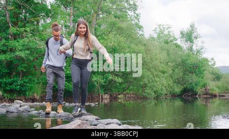 Young Couple Crossing River On Stepping Stones Hiking In UK Lake District Stock Photo
