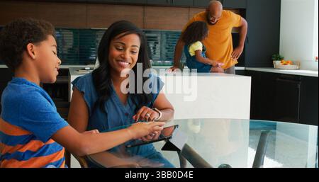 African American Parents Helping Children Using Digital Devices In Kitchen Stock Photo