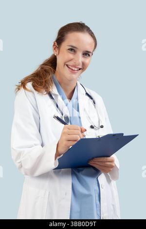 Smiling female doctor in a white coat writing a prescription or patient notes on a clipboard. Medical professional with stethoscope isolated on light Stock Photo