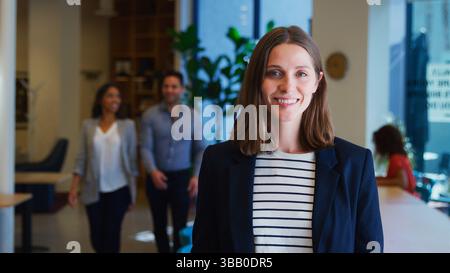 Portrait Of Businesswoman Standing In Busy Modern Open Plan Office With Colleagues In Background Stock Photo