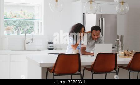 Loving Couple Wearing Pyjamas Standing In Kitchen Working From Home On Laptop Stock Photo