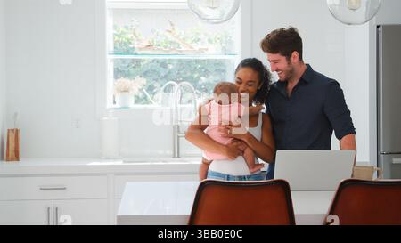 Family With Baby Daughter In Kitchen Using Laptop On Counter Stock Photo
