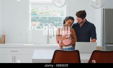 Family With Baby Daughter In Kitchen Using Laptop On Counter Stock Photo