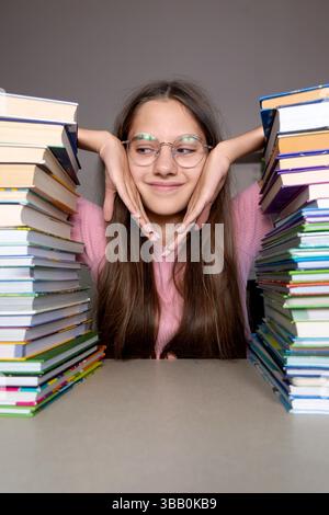 Thoughtful student sitting between stacks of books. A young student wearing glasses sits with her chin resting on her hands between two tall stacks of Stock Photo