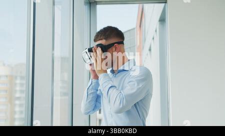 Man using VR headset near large window. A man in a blue shirt stands near a large window while using a virtual reality headset. Stock Photo