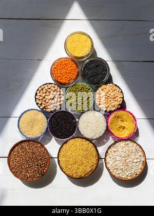 Assorted Grains and Legumes in Bowls Arranged in Pyramid Shape on White Wooden Background. Colorful grains, legumes, and pasta arranged in a pyramid Stock Photo