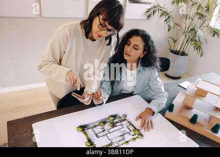 Two women collaborating in an architectural office, one showing plans and using a smartphone for discussion, inspiring teamwork. Stock Photo