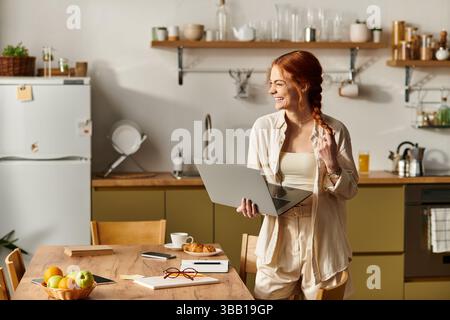 Brightly lit kitchen showcases a cheerful red haired woman engaged in creative work while smiling. Stock Photo