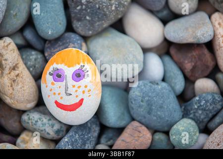 White stone with a painted positive face on a background of gray sea pebbles.Concept of creativity. Stock Photo