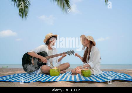 Young Asian couple enjoying a tropical beach moment with guitar music and coconut drinks on a striped picnic mat. The woman claps along while the man Stock Photo