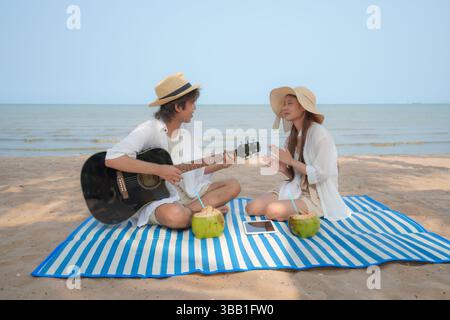 Young Asian couple enjoying a tropical beach moment with guitar music and coconut drinks on a striped picnic mat. The woman claps along while the man Stock Photo