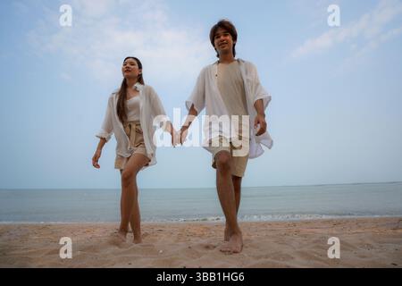 Young Asian couple enjoying a relaxing walk along the sandy beach under a clear blue sky, dressed in matching light casual outfits, symbolizing romant Stock Photo