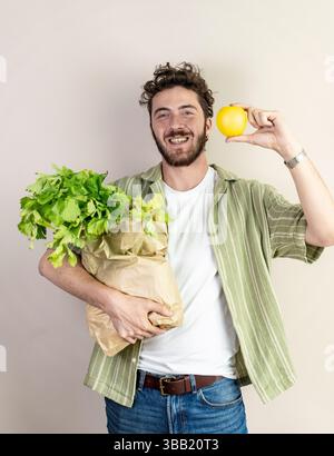 Portrait of a smiling millennial man showing a paper grocery bag full of leafy greens while raising a yellow apple. Concept of healthy eating, sustain Stock Photo