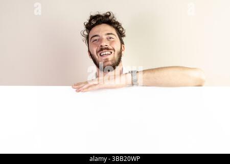 Cheerful millennial male leaning on a large horizontal poster board with empty space for text or graphic design. Concept for announcement, marketing m Stock Photo