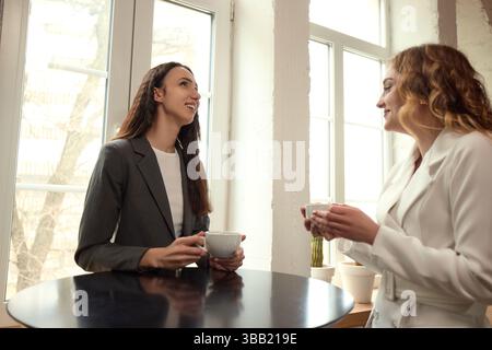 Colleagues converse near high table in modern office during break, holding coffee cups, smiling. Stock Photo
