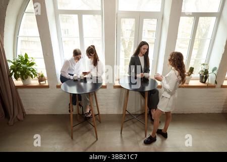 Colleagues gather near high tables in modern office during break, some check phones, holding coffee cups. Stock Photo